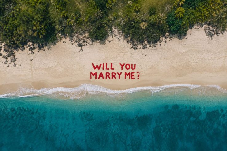 A flower-petal message reading Will You Marry Me on a beach, seen from the air
